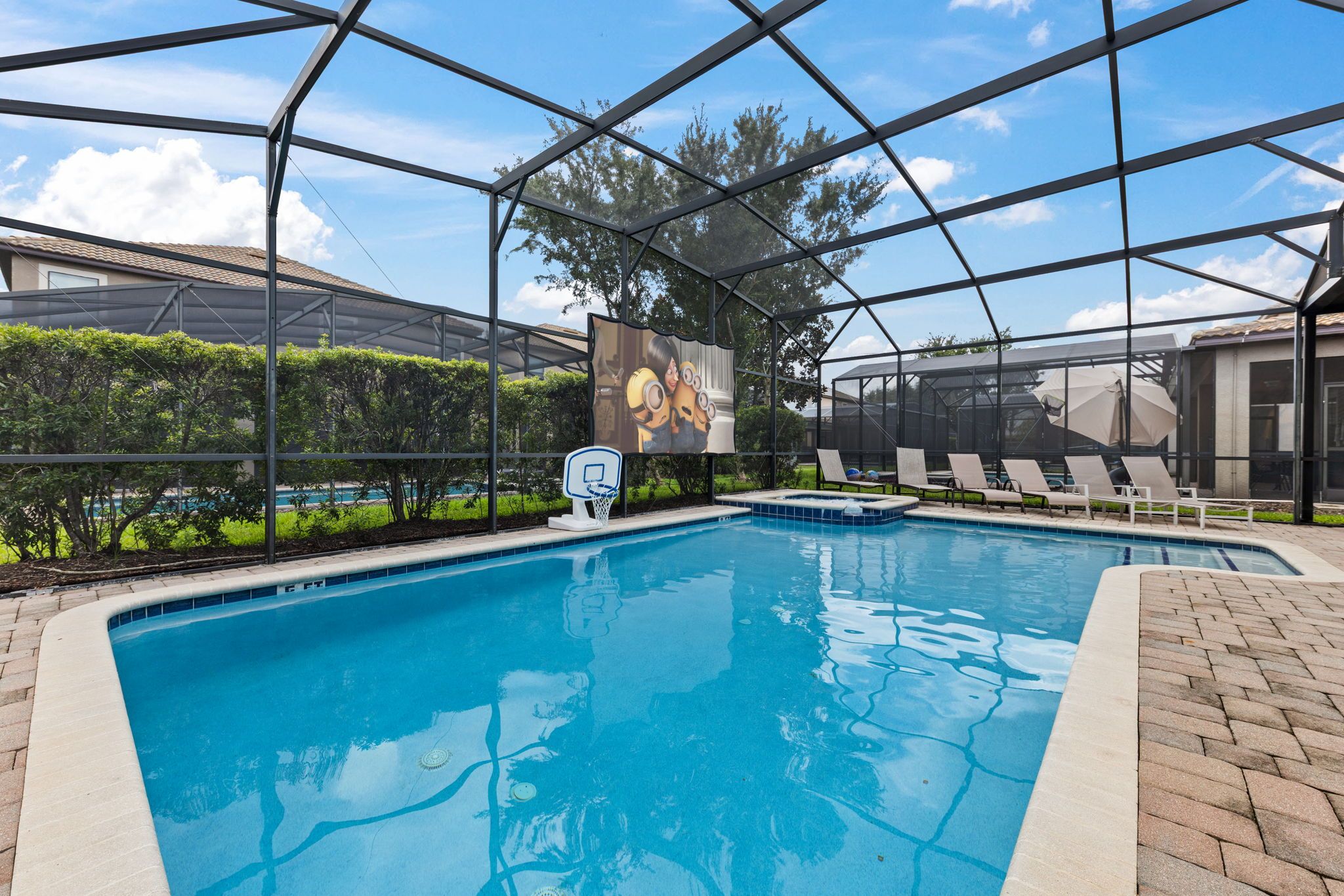 A swimming pool with a basketball hoop, lounge chairs, and a large screen displaying animated characters.