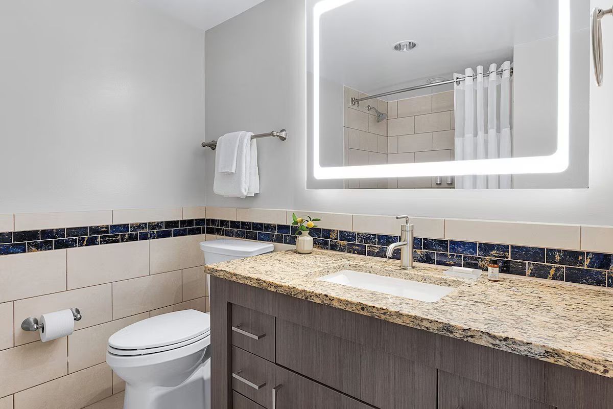 A modern bathroom with a white toilet, a sink with a granite countertop, and a mirror with integrated lighting.