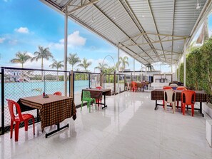 Een terras buiten met tafels en stoelen, uitzicht op zee, en een overdekt gedeelte met een dak.