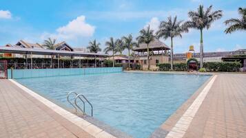 A swimming pool with a clear blue water surface, surrounded by a tiled deck, palm trees, and a building with a thatched roof.