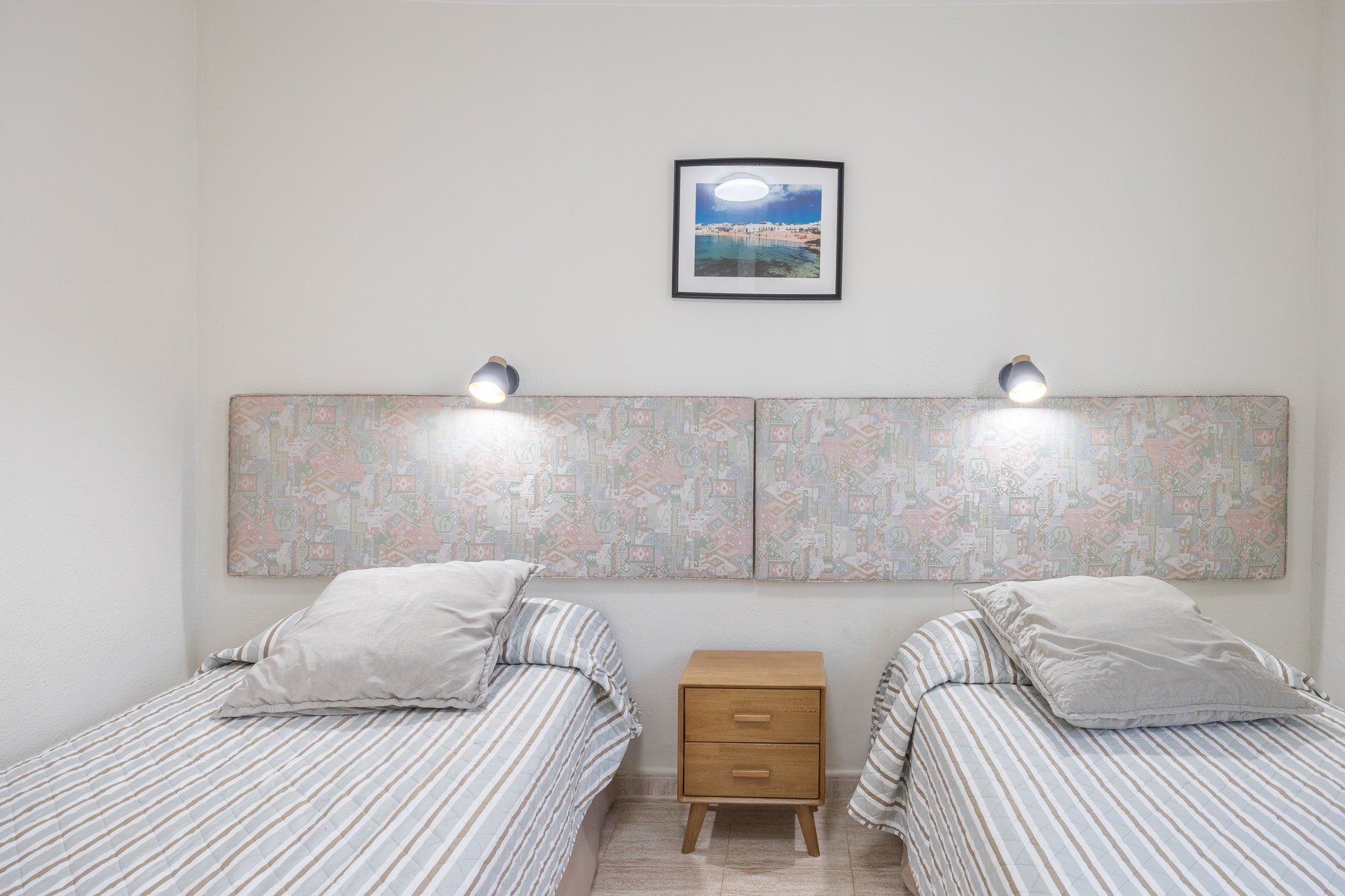 Un dormitorio con dos camas, una mesita de noche, un cuadro en la pared y dos lámparas de pared.