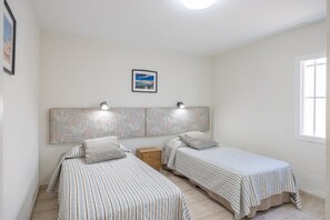 A bedroom with two single beds, a headboard with a decorative pattern, a nightstand, and a framed picture on the wall. - GuestReady - Los Alisios Corralejo 39 (La Oliva)