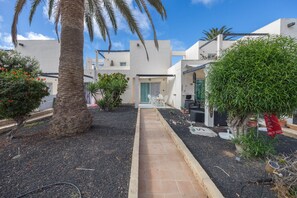 A pathway leading to a building with a door, surrounded by greenery and a palm tree. - GuestReady - Los Alisios Corralejo 39 (La Oliva)