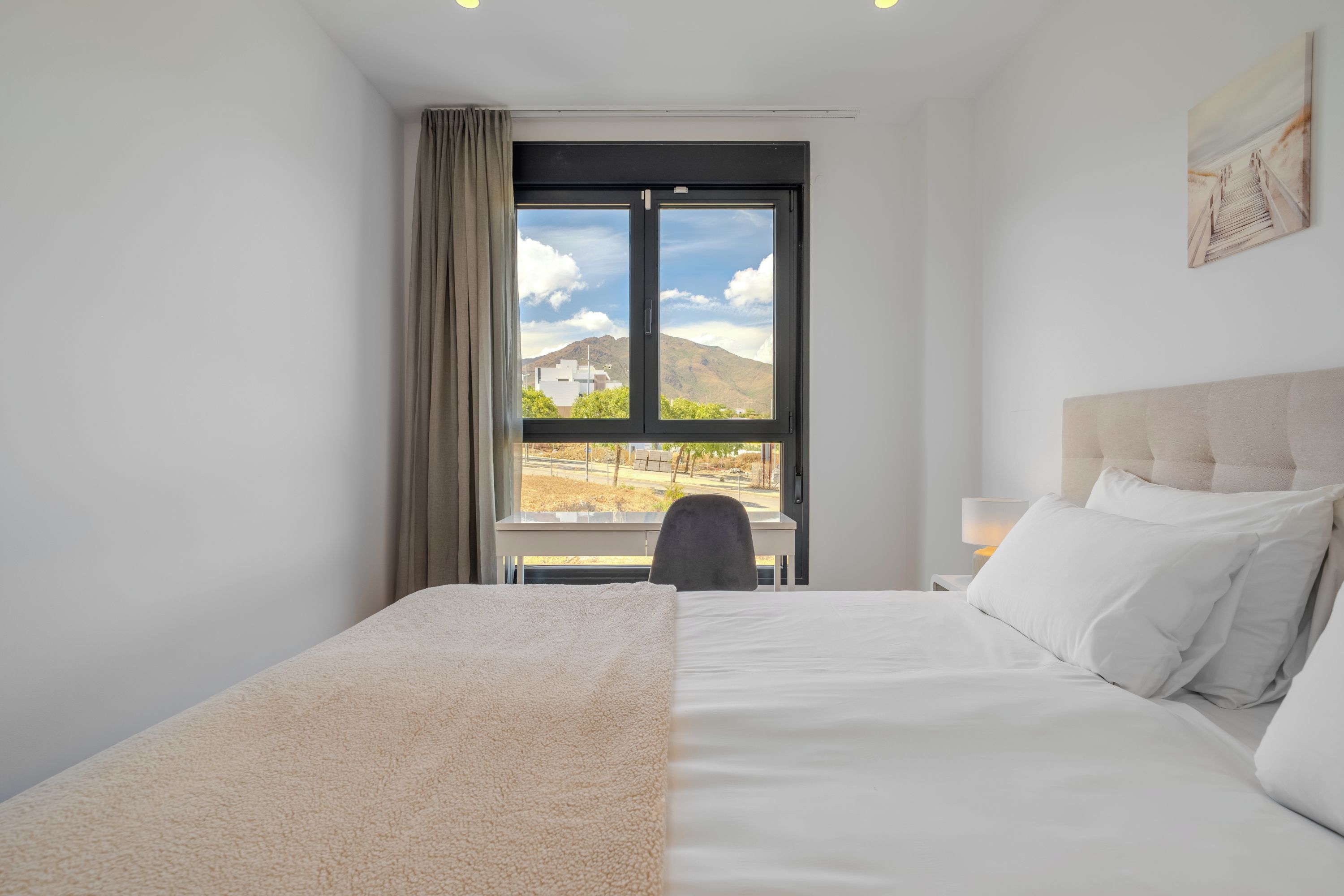 A hotel room with a bed, a chair, a desk, a lamp, and a window with a view of a mountain and buildings.