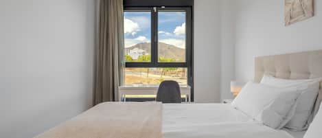 Een hotelkamer met een bed, een stoel, een bureau, een lamp en een raam met uitzicht op een berg en gebouwen.