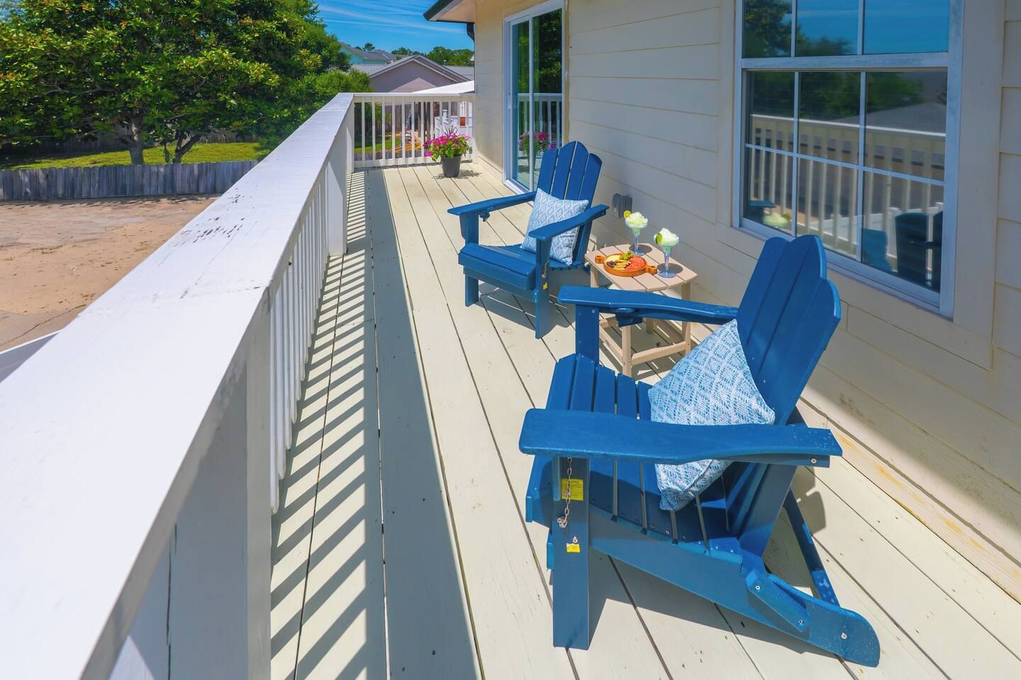 A wooden deck with blue Adirondack chairs and a small table with drinks, overlooking a backyard.