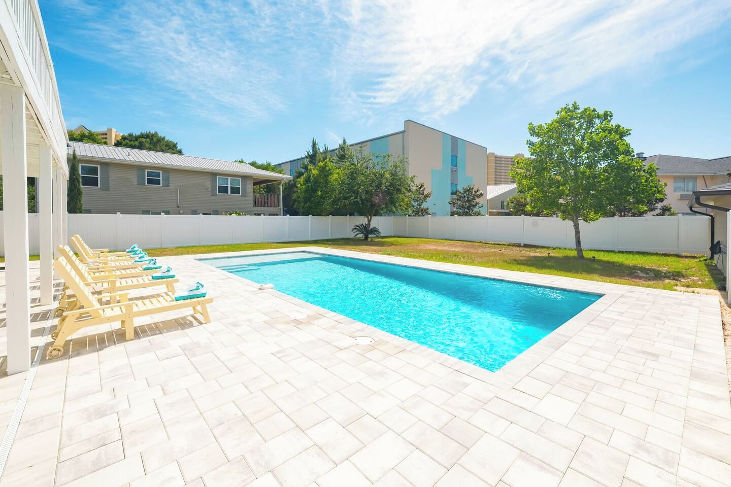 Une piscine rectangulaire, bordée d’une terrasse carrelée, entourée d’une clôture blanche, et une rangée de chaises longues en bois disposées sur un des côtés.