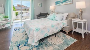 A bedroom with a bed, nightstands, a lamp, a painting, and a view through a window. - 1/2 Block to the Beach, West End | Balcony+Pool (Panama City Beach)