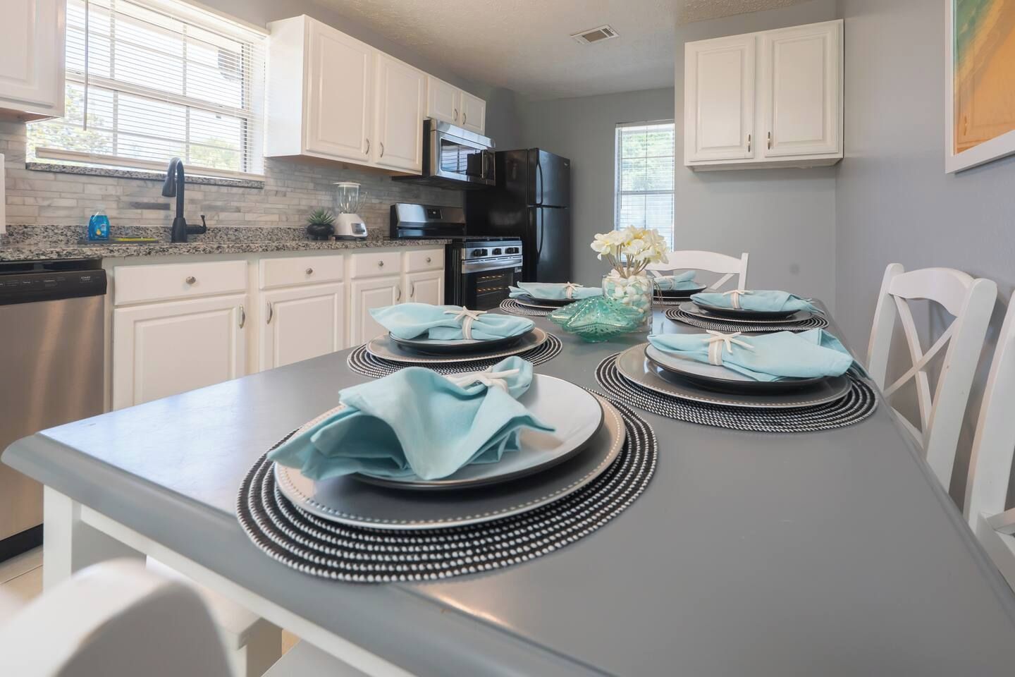 A modern kitchen with white cabinets, a stainless steel refrigerator, and a dining table set with plates and napkins.