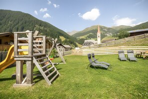 A playground with a slide and climbing structures, surrounded by a grassy area with benches and a view of a mountain.