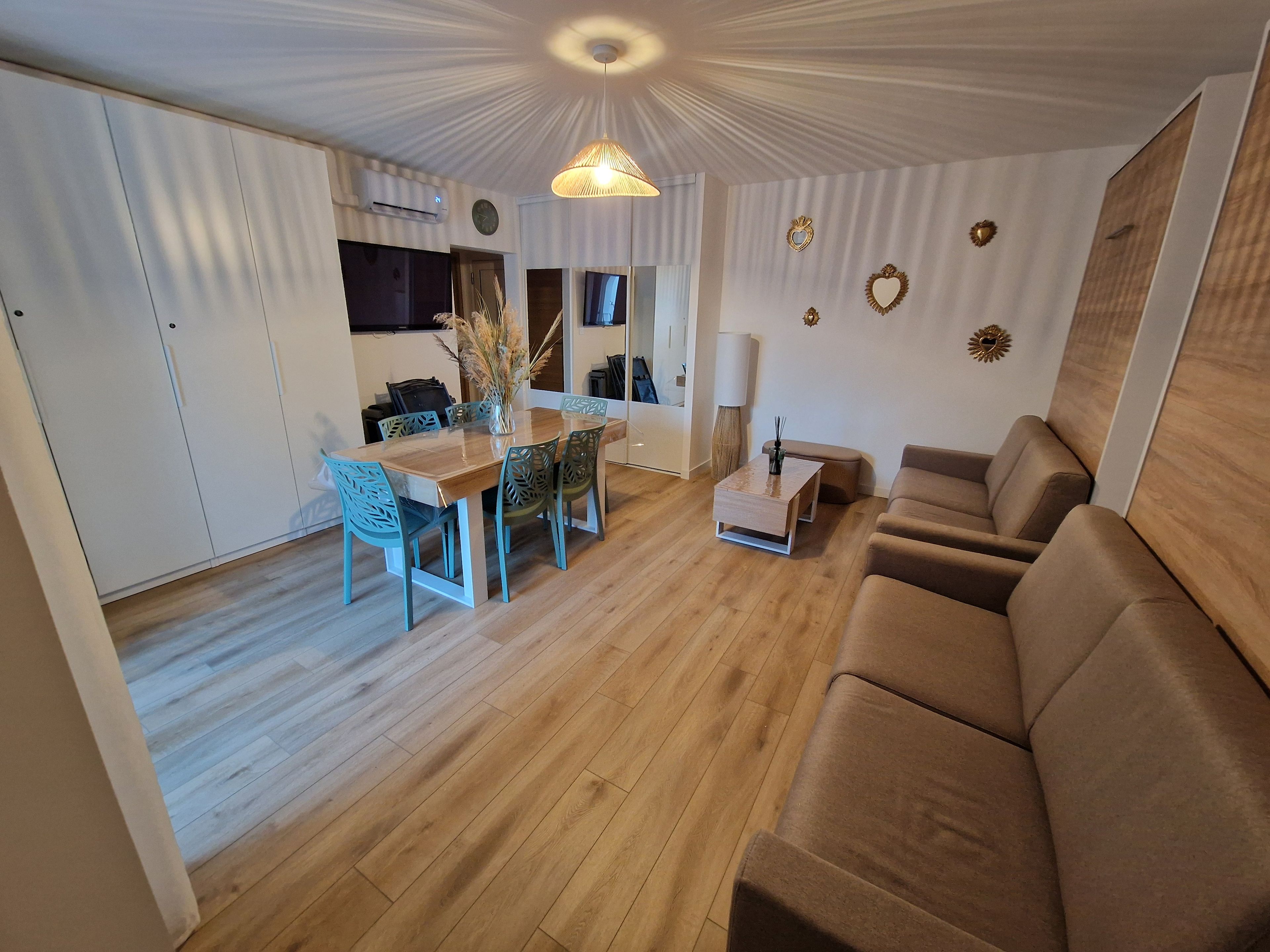 Ein modernes Wohnzimmer mit Holzboden, einem braunen Sofa, einem Esstisch mit Stühlen und einer Küchenzeile im Hintergrund.