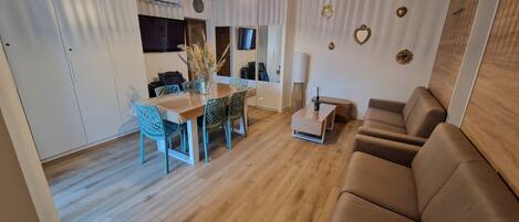 Ein modernes Wohnzimmer mit Holzboden, einem braunen Sofa, einem Esstisch mit Stühlen und einer Küchenzeile im Hintergrund.