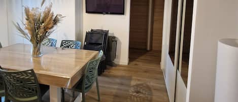 Un coin repas avec une table en bois, des chaises, un vase contenant des plantes séchées, un téléviseur à écran plat fixé au mur et une porte donnant sur une autre pièce.