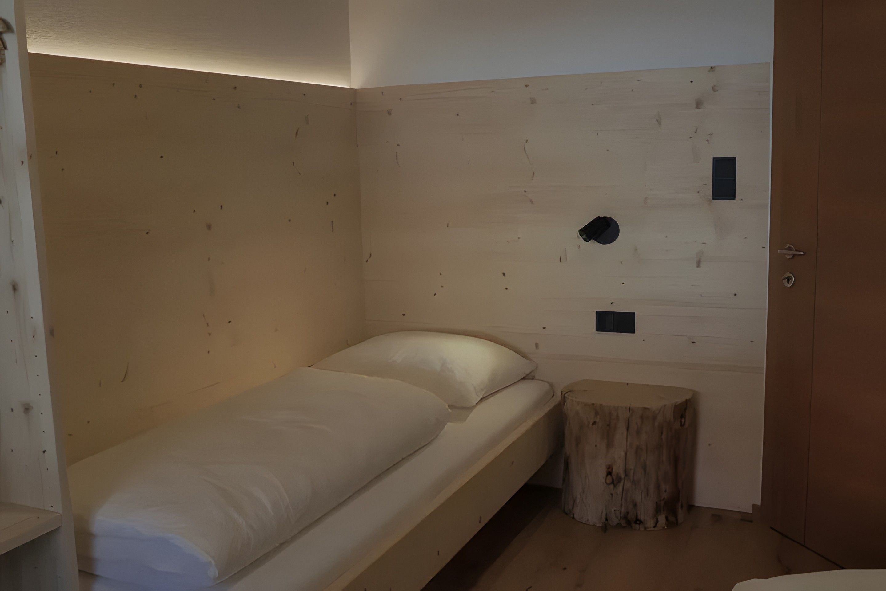 Ein kleines Zimmer mit einem Einzelbett, einem Holzkopfstück, einem kleinen Hocker und einer Tür.
