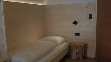 Ein kleines Zimmer mit einem Einzelbett, einem Holzkopfstück, einem kleinen Hocker und einer Tür.