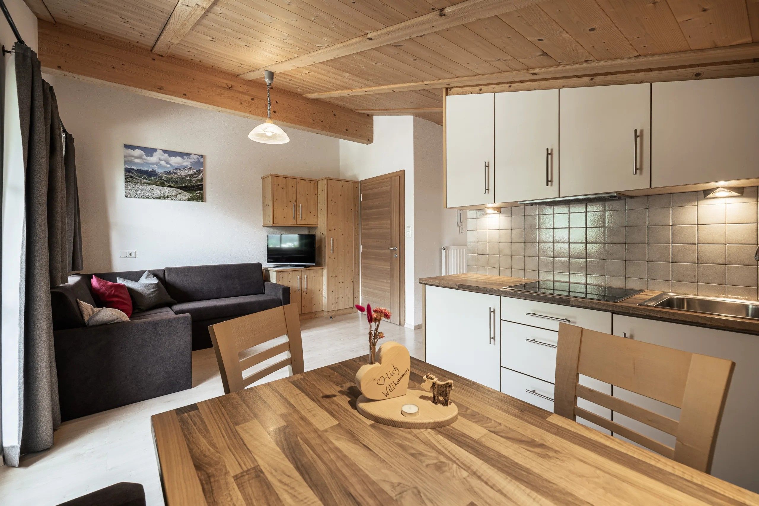 Eine moderne Küche mit weißen Schränken, einem Holztisch im Essbereich und einem Fernseher im Wohnzimmer.