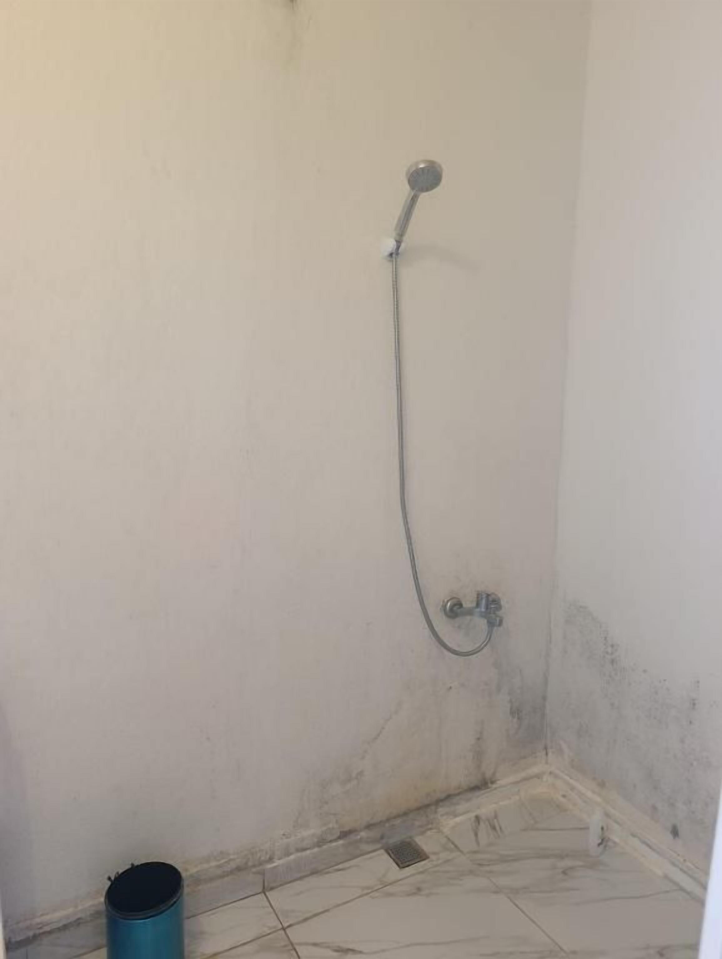 Un baño pequeño con poca luz, ducha, desagüe visible y un cubo de basura azul.