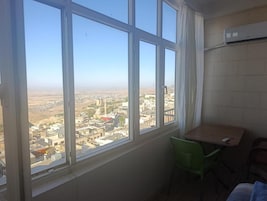 Quarto com uma janela grande com vista para a paisagem urbana, uma mesa pequena e uma cadeira verde.