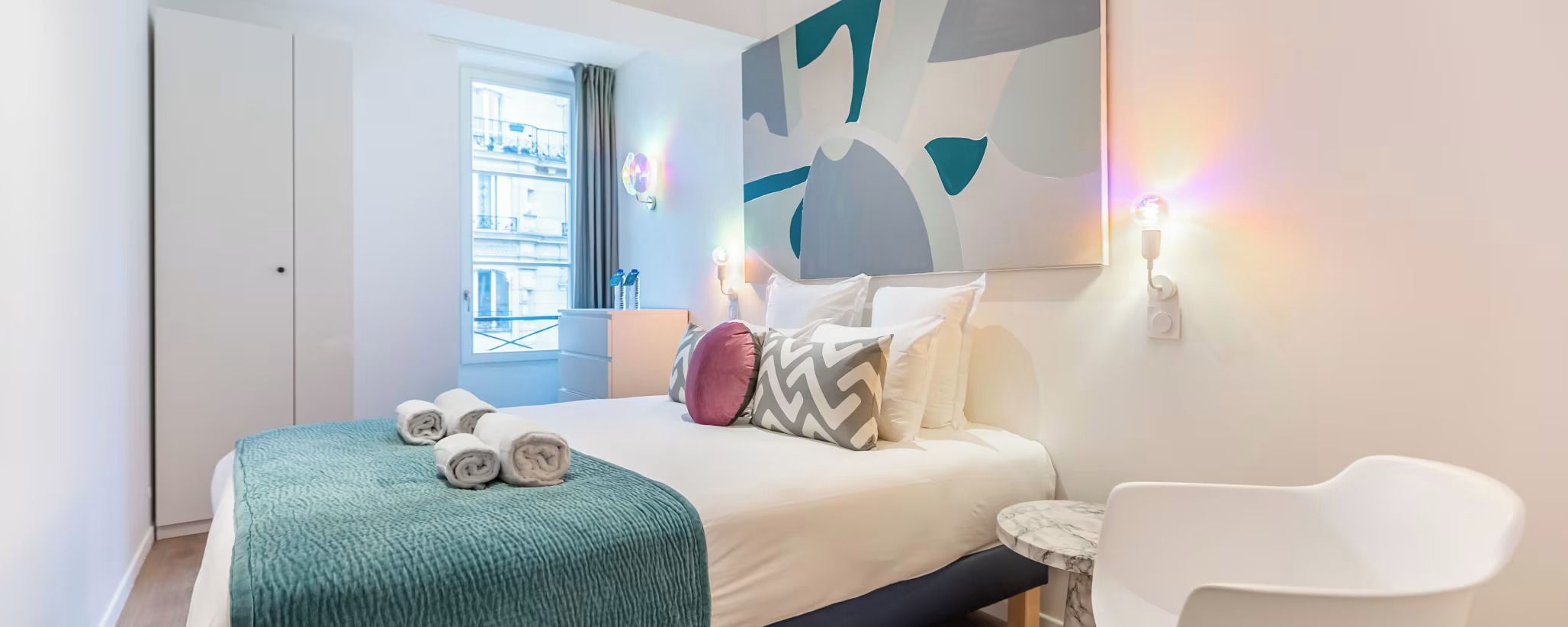 A hotel room with a bed, a small round table, a chair, a window with a view of buildings, and a wall with abstract art.