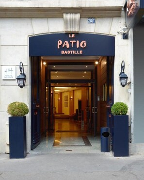 Der Eingang zum Hotel Le Patio Bastille mit einer dunklen Fassade, einem einladenden Bogen und einem gut beleuchteten Innenraum, der durch die Tür sichtbar ist.