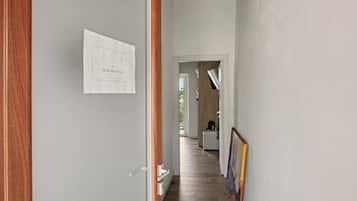 Een gang met een deur aan de linkerkant, een lijst met een bord op de muur, en een keuken in de verte.