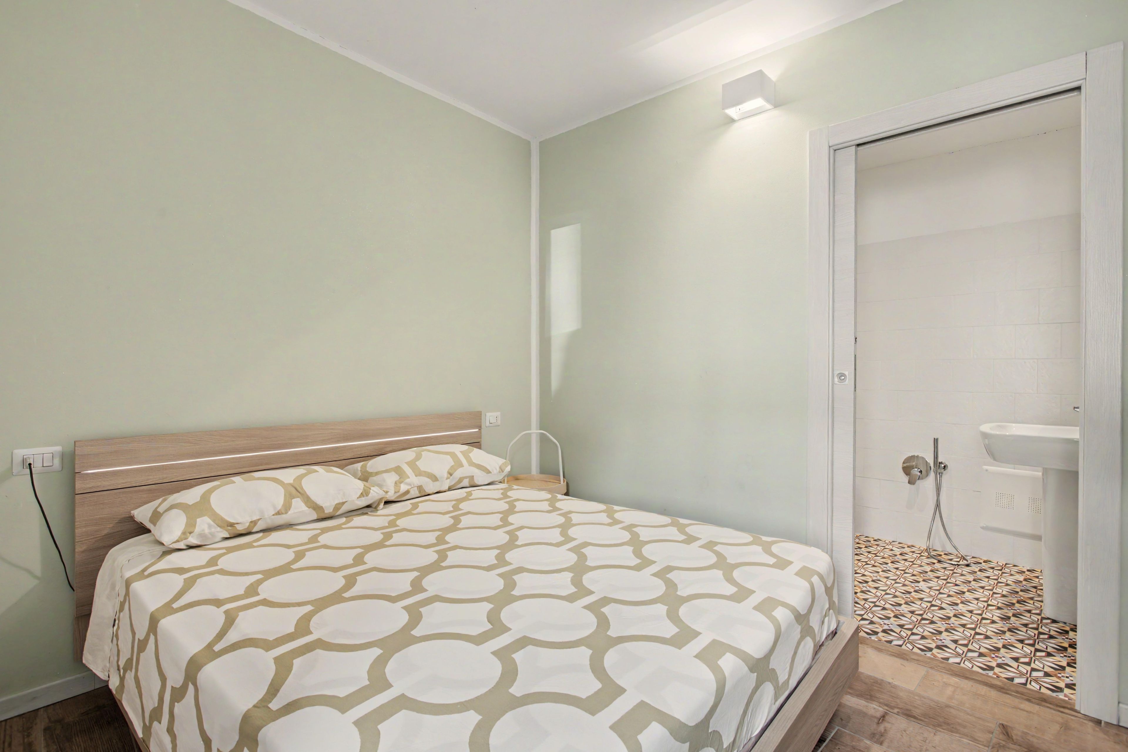 A bedroom with a bed, a bedside table, a wall-mounted light, and a bathroom with a shower and sink.