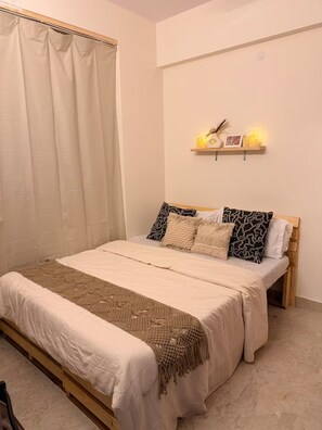 A bedroom with a bed, pillows, a bedside table, and a wall-mounted shelf with decorative items. - The golden hour by Elora Homes: 2BHK in Arekere (Bengaluru)