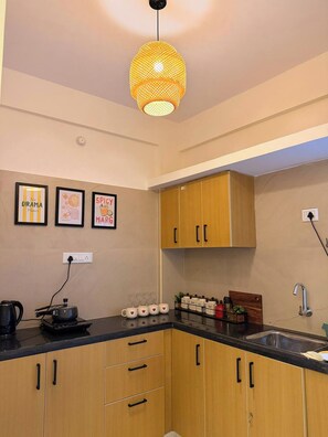 A kitchen with wooden cabinets, a black countertop, a hanging light fixture, and two wall-mounted signs. - The golden hour by Elora Homes: 2BHK in Arekere (Bengaluru)