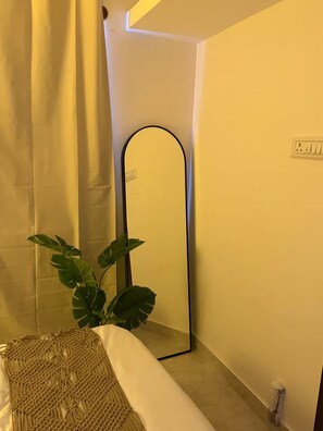 Een kamer met een bed, een plant, een spiegel en een lichtschakelaar aan de muur.