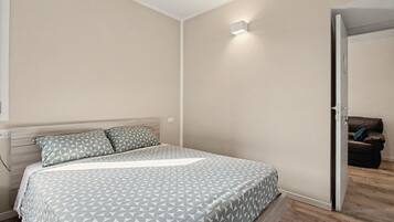 A bedroom with a bed, a bedside table, a wall-mounted light, and a door leading to another room.