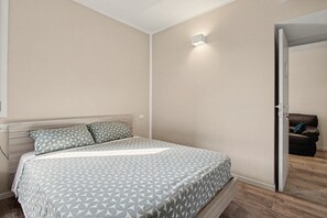 Un dormitorio con cama, mesita de noche, luz empotrada en la pared y una puerta que da a otra habitación.