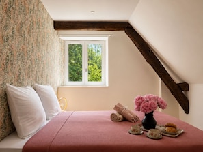 A bedroom with a large window, a bed with a pink bedspread, a vase with flowers, and a tray with pastries and cups.
