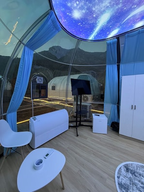 Una habitación moderna y cerrada con techo curvo que muestra un cielo estrellado, un área de estar con una mesa y vistas a una estación de tren.