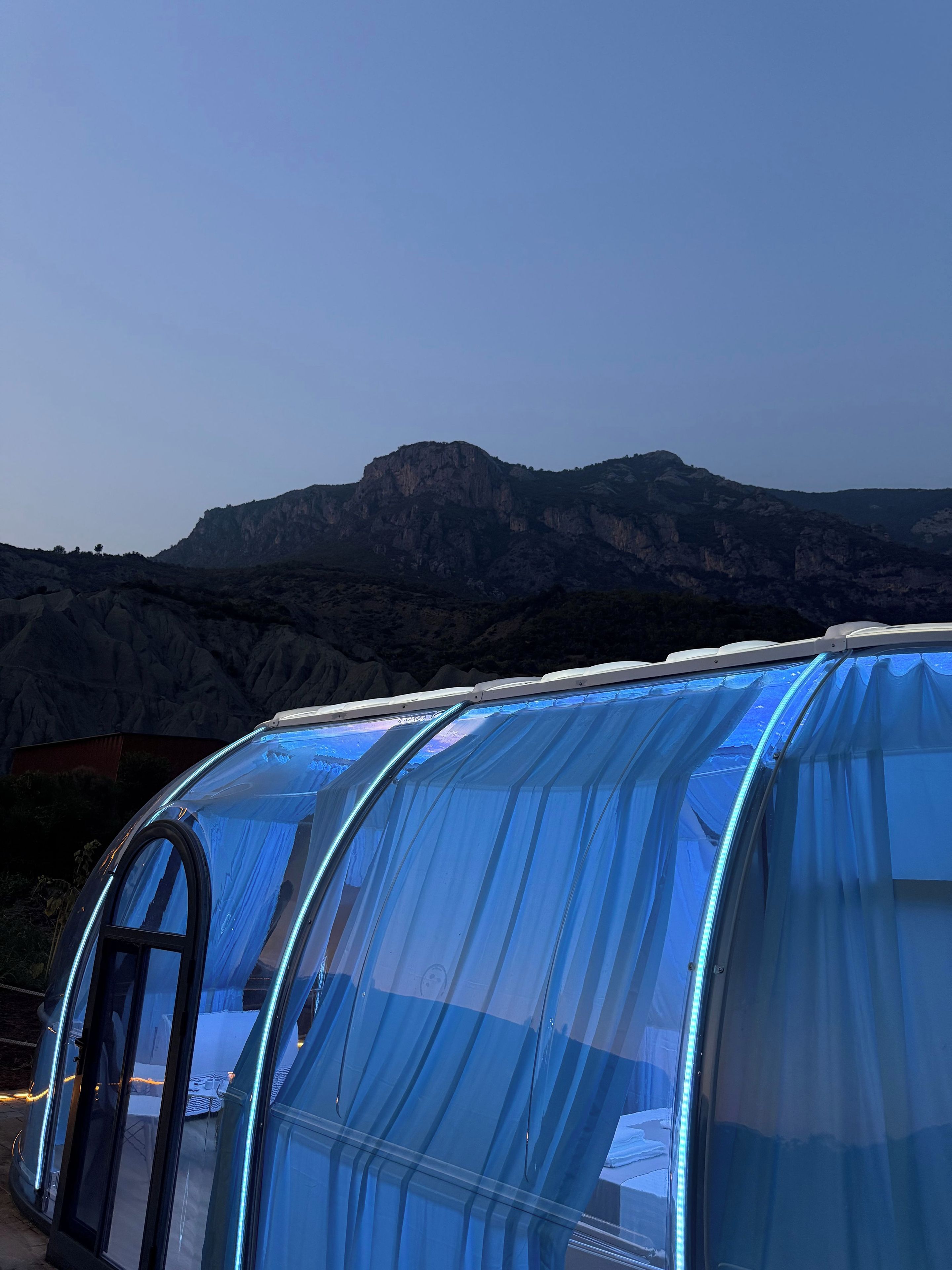 Una estructura transparente con techo curvo, posiblemente un invernadero o una carpa, con montañas al fondo.