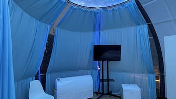 Una habitación tipo tienda de campaña con techo estrellado, televisor de pantalla plana, sofá blanco, una mesita y una silla blanca.