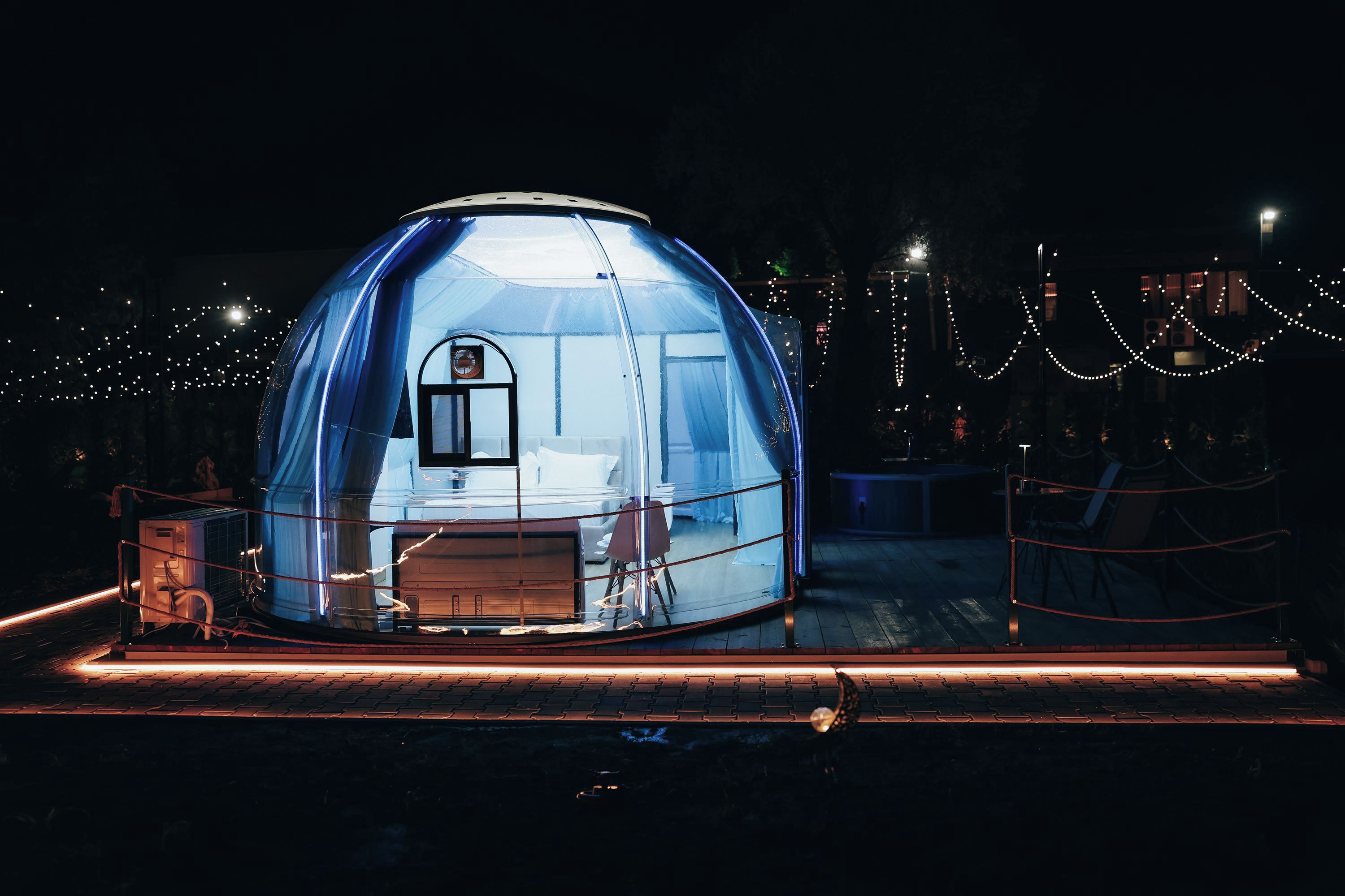 Une structure en forme de dôme, comprenant un lit et une petite table, entourée d’une clôture et illuminée de guirlandes lumineuses.