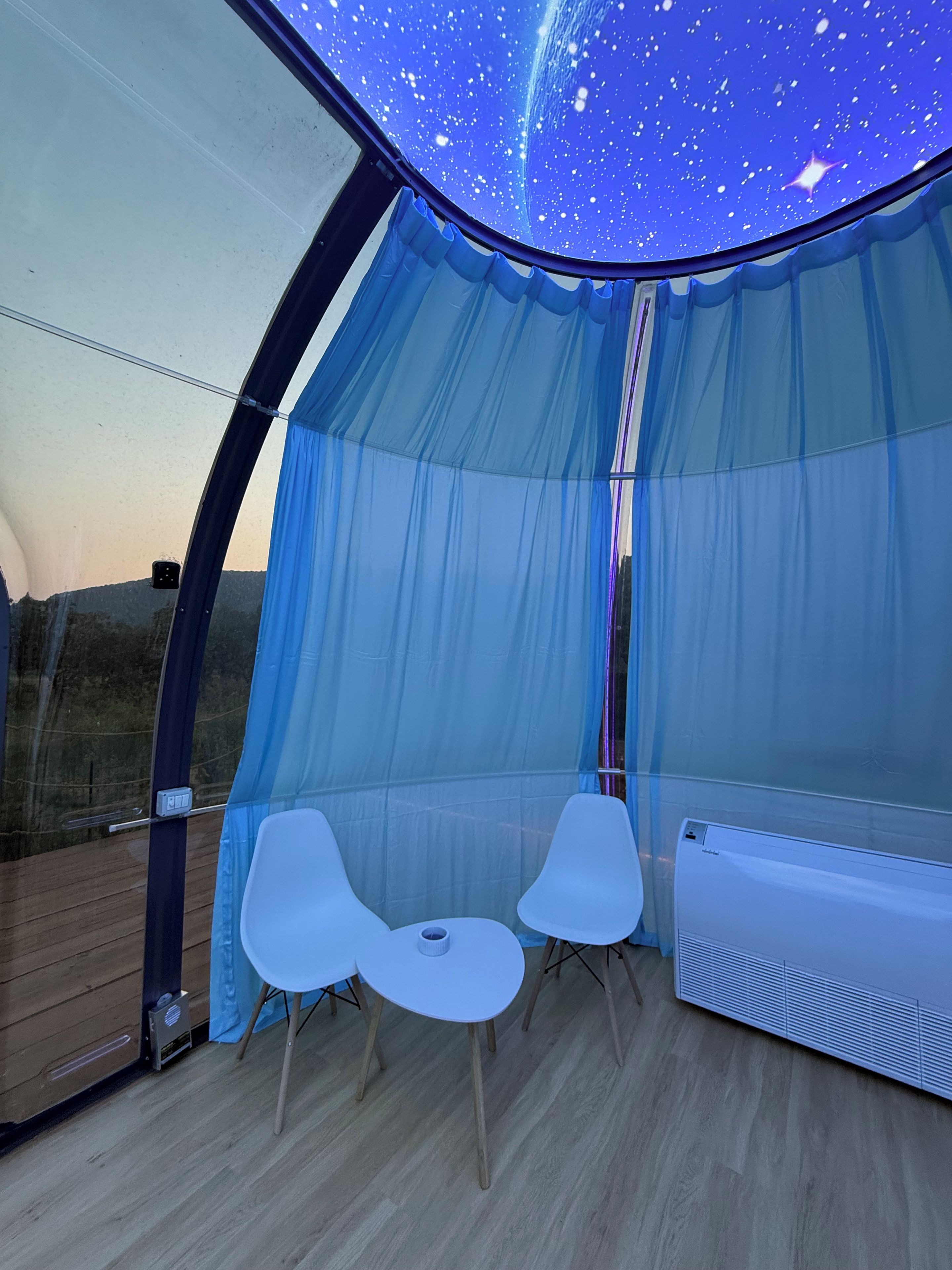 Una habitación con techo que simula un cielo estrellado, dos sillas blancas y un equipo de aire acondicionado blanco.