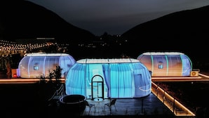 Three yurts with a semi-transparent dome structure, each with a small window and a door, situated on a wooden deck with a metal railing.