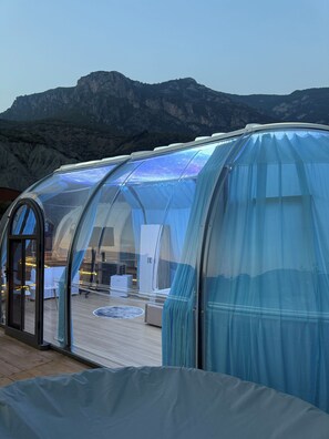 A transparent dome structure with a wooden deck and seating area, set against a mountainous backdrop. - Magas Bubble Houses (Dajt)