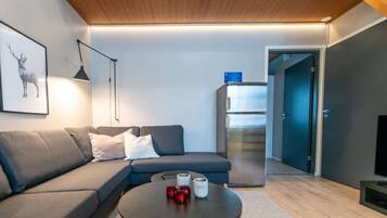 Ein modernes Wohnzimmer mit einem Ecksofa, einem kleinen runden Tisch mit Kerzen, einer Wandleuchte und einem Kühlschrank.