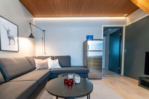 A modern living room with a sectional sofa, a small round table with candles, a wall-mounted lamp, and a refrigerator.
