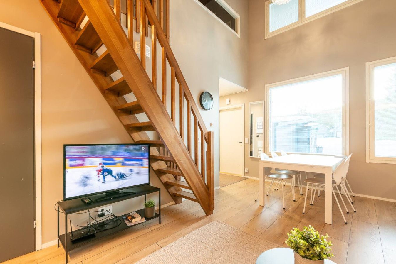 A modern living room with a wooden staircase, a flat-screen TV, a dining area with a white table and chairs, and large windows.