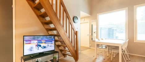 A modern living room with a wooden staircase, a flat-screen TV, a dining area with a white table and chairs, and large windows.