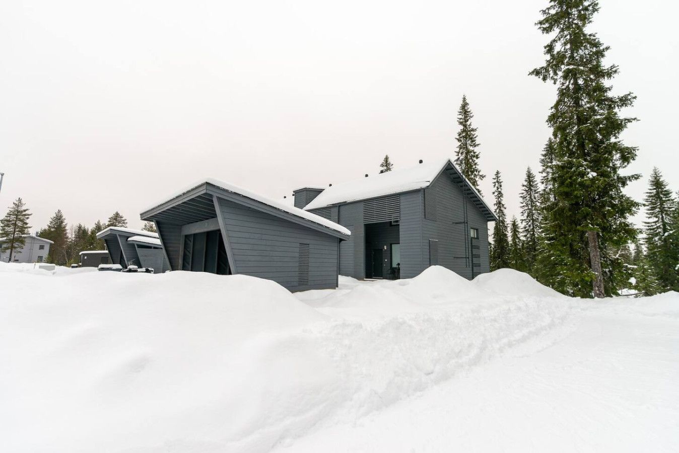 A modern building with a sloped roof, surrounded by snow and tall evergreen trees.