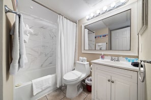 A bathroom with a bathtub, toilet, sink, and a mirror with lights above. - Cute condo on Seawall across from beach (Galveston)