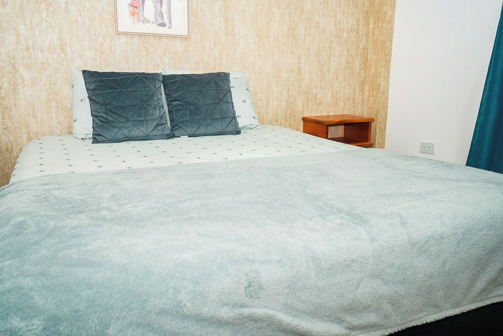 A bed with a quilted headboard, a small wooden nightstand, and a wall-mounted picture.
