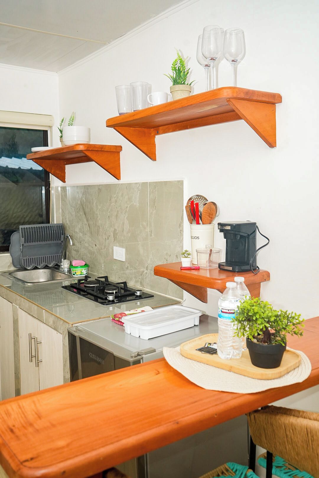 Una cocina moderna con estantes de madera, estufa a gas, lavamanos y una zona de comedor con una silla.
