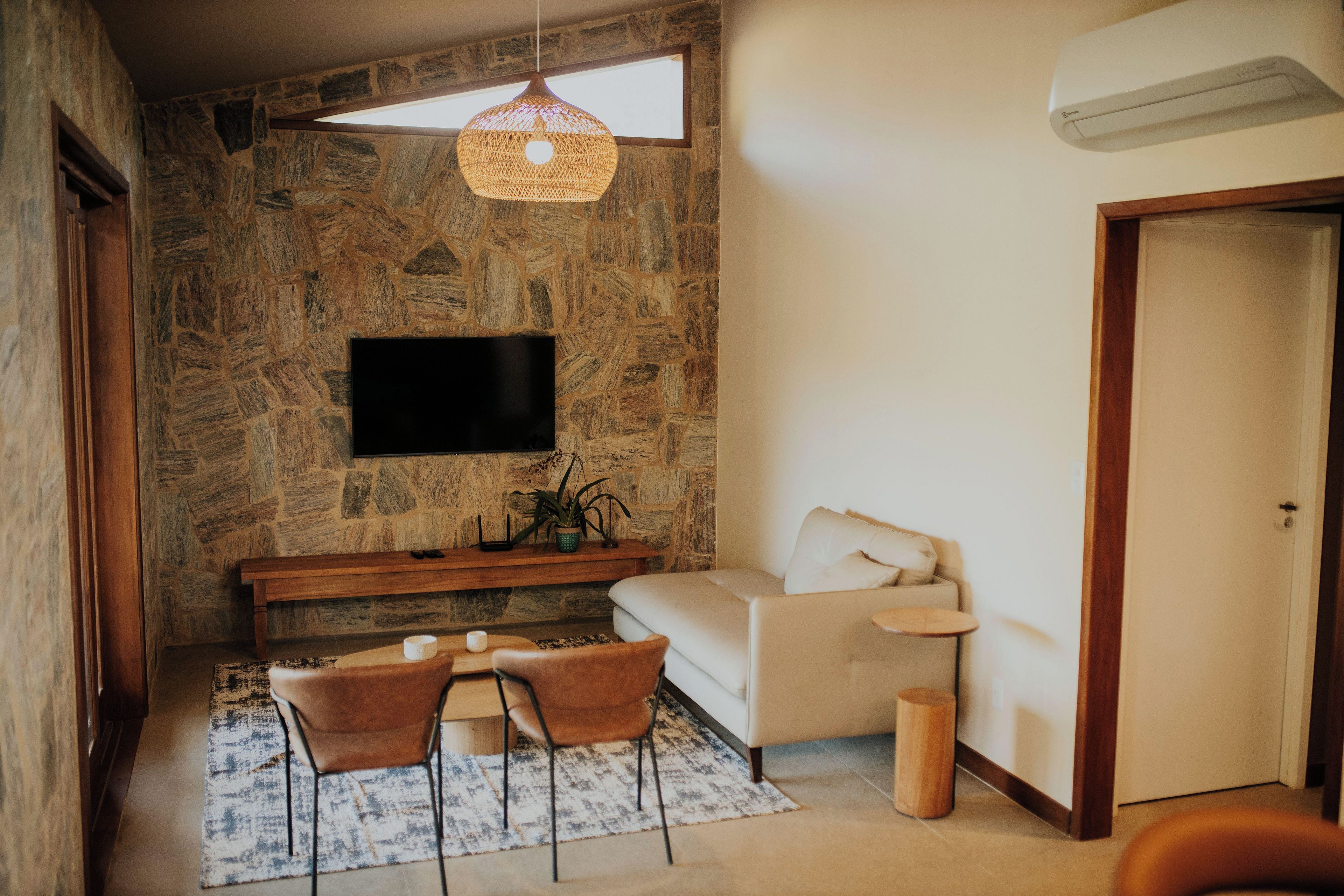 A living room with a stone wall, a flat-screen TV, a sofa, a coffee table, two chairs, a hanging light fixture, and a small side table.