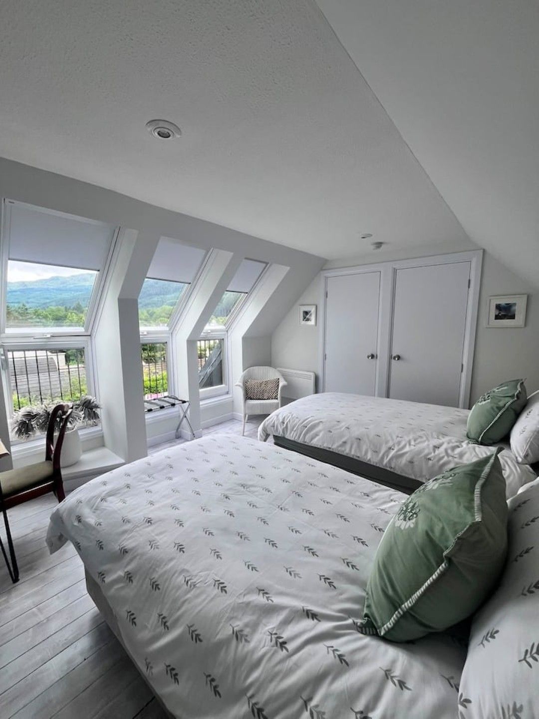 Une chambre avec un grand lit, deux armoires blanches, une fenêtre donnant sur un paysage verdoyant et un fauteuil.