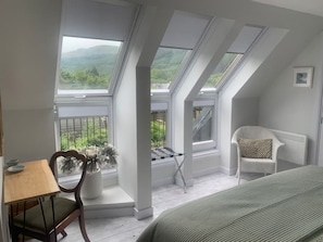 A room with a sloped ceiling, a view of greenery outside, a white chair, a small table, and a bed.